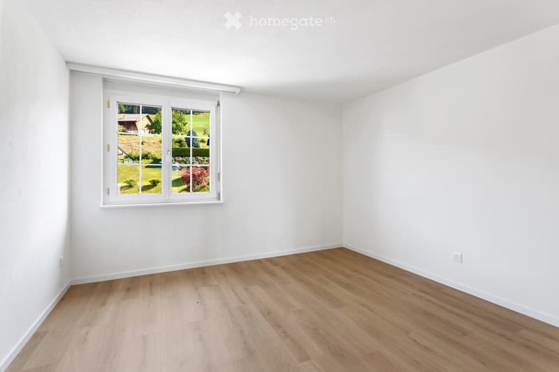 Empty room with white walls and light wooden flooring, featuring a large window with white frame showing a view of a garden and house outside.