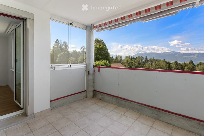 Balcony with tiled floor, white walls, red trim, large windows offering a scenic mountain view.