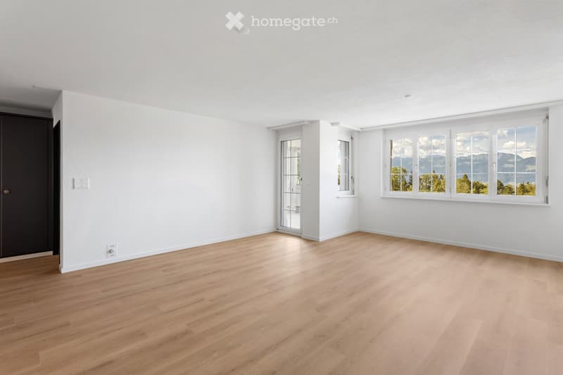 Empty room with light wooden flooring, white walls, and large windows offering a scenic view of mountains and trees.