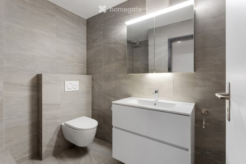 Modern bathroom with grey tiled walls, white sink with built-in cabinet, toilet, large mirror with integrated lighting, and a sleek faucet.