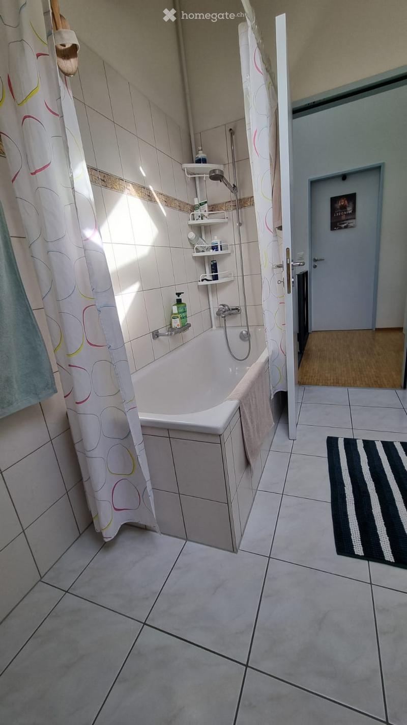 Bathroom with a bathtub, shower curtain, towel, and bathroom shelf with bottles. The floor is tiled, and there is a black and white striped rug near the door.