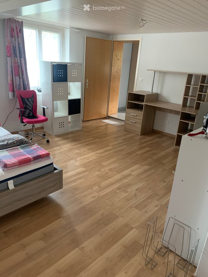 Empty bedroom with wooden floor, white walls, double bed, desk, chair, wooden storage units, window with curtains, rug by the door, electrical outlet and light fixture.
