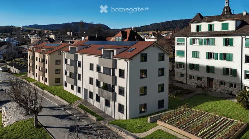 Modern multi-story residential building with solar panels on the roof, surrounded by a landscaped garden area with raised beds and pathways.