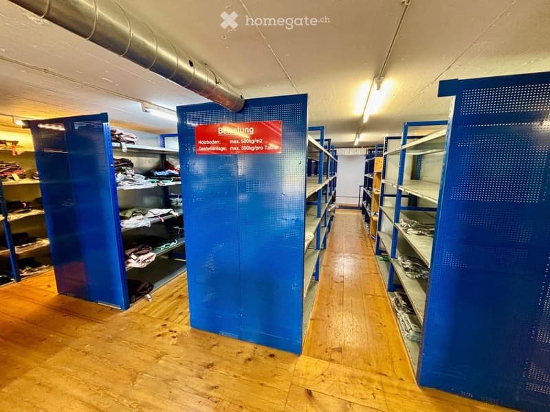 A long, narrow hallway with wooden flooring and blue shelving units on both sides, containing folded clothes and other stored items. The hallway is well-lit with fluorescent lighting and has a sign indicating weight limits for storage.