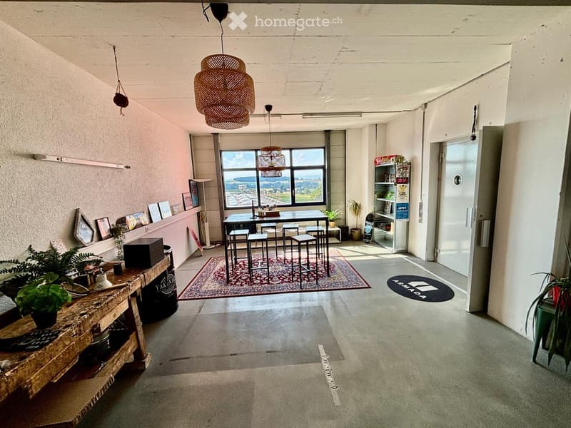 Open-plan space with a dining area, bar counter, and shelves. Features a large window with a scenic view, wicker pendant lights, and potted plants.