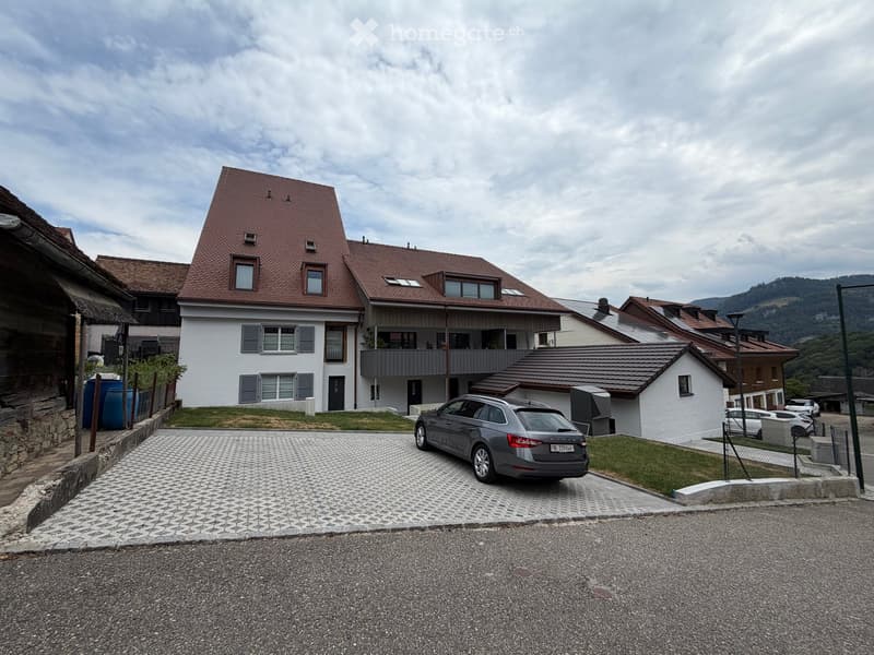 Two-story house with a red-tiled roof, white walls, multiple windows, and a balcony on the upper floor. A grey car is parked in the driveway.