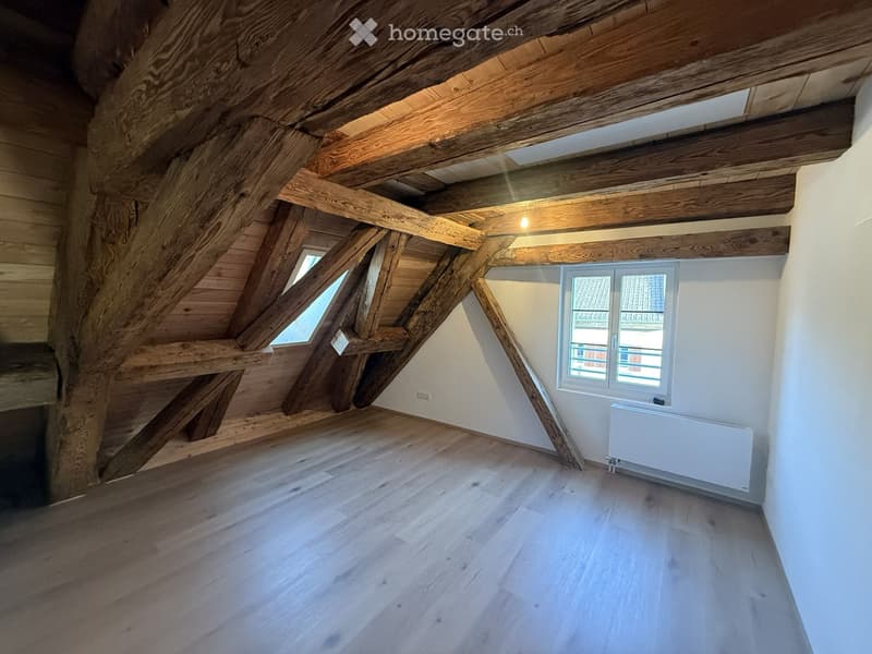 Empty attic space with wooden beams, wooden floor, white walls, and two windows, one small skylight and one larger window with a view of the roof.