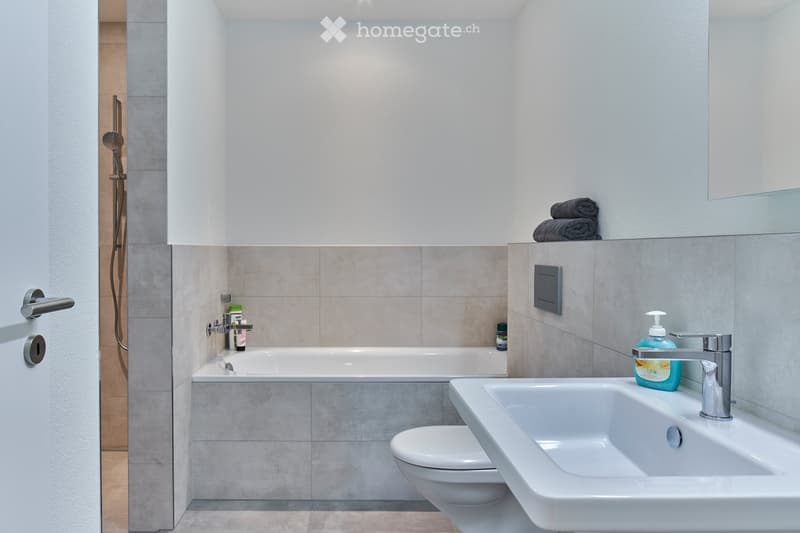 Modern bathroom with a bathtub, sink, toilet, and shower. Features a sleek design with light-colored tiles and a large mirror above the sink.