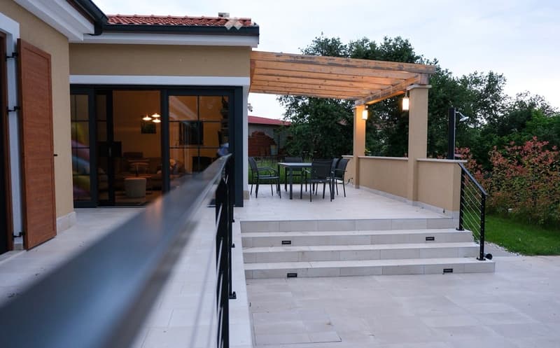 Balcony with steps leading to a covered wooden pergola, featuring a dining set with chairs and a table, surrounded by greenery and a modern house.