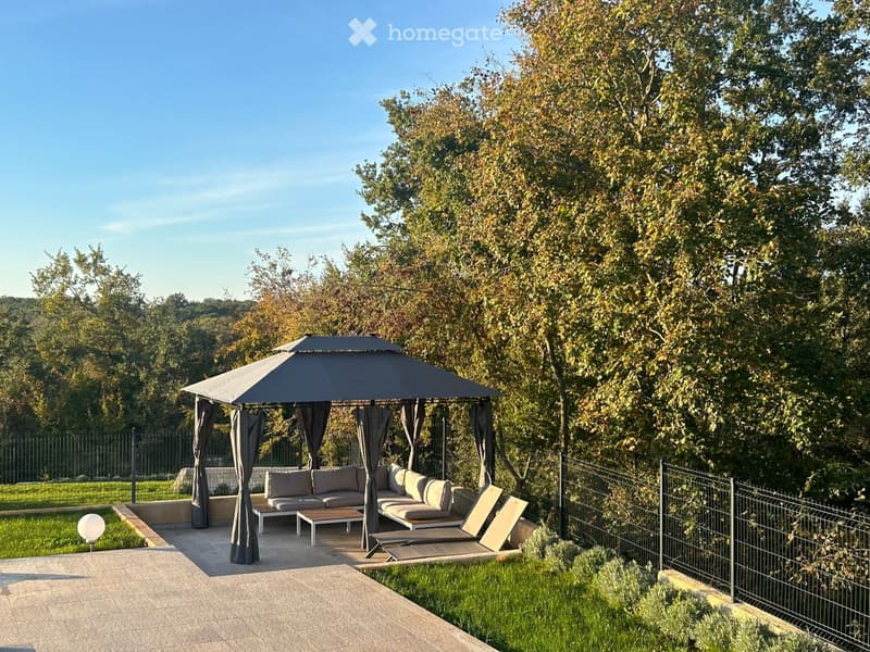 A well-maintained garden area features a large patio with a covered seating area, including a sofa, chairs, and a table, surrounded by lush greenery and a clear blue sky.