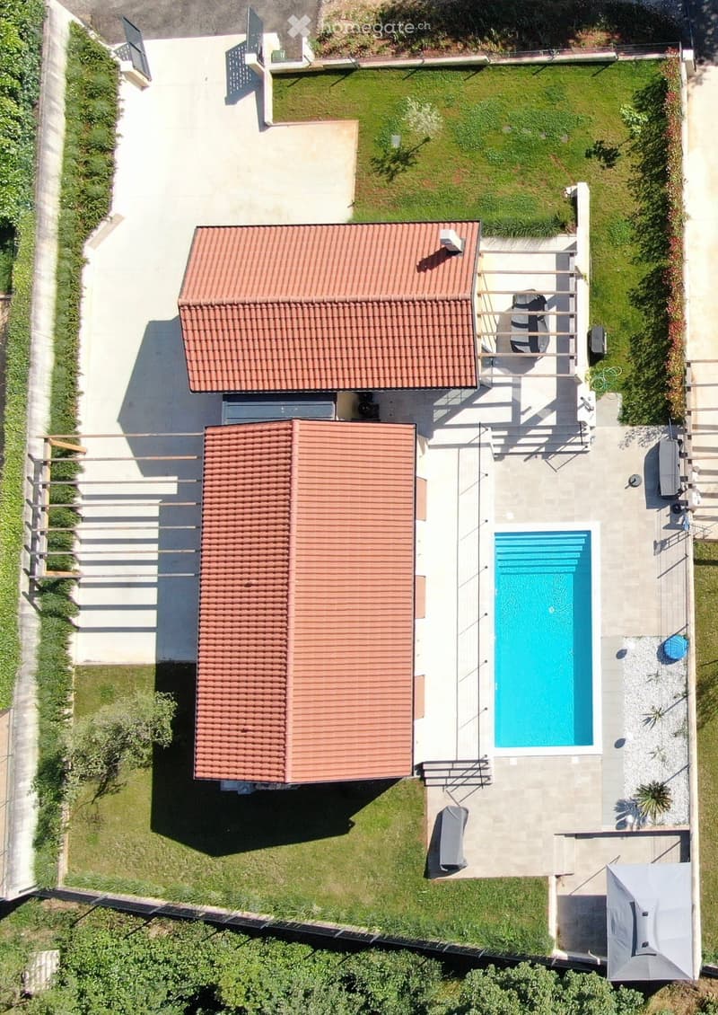 Aerial view of a two-story house with a red-tiled roof, surrounded by a well-maintained garden and a swimming pool.