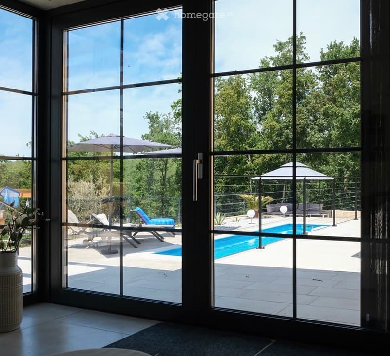 Bright room with large glass doors opening to a patio with a swimming pool, lounge chairs, and umbrellas. A vase with flowers is placed on a table near the glass doors.