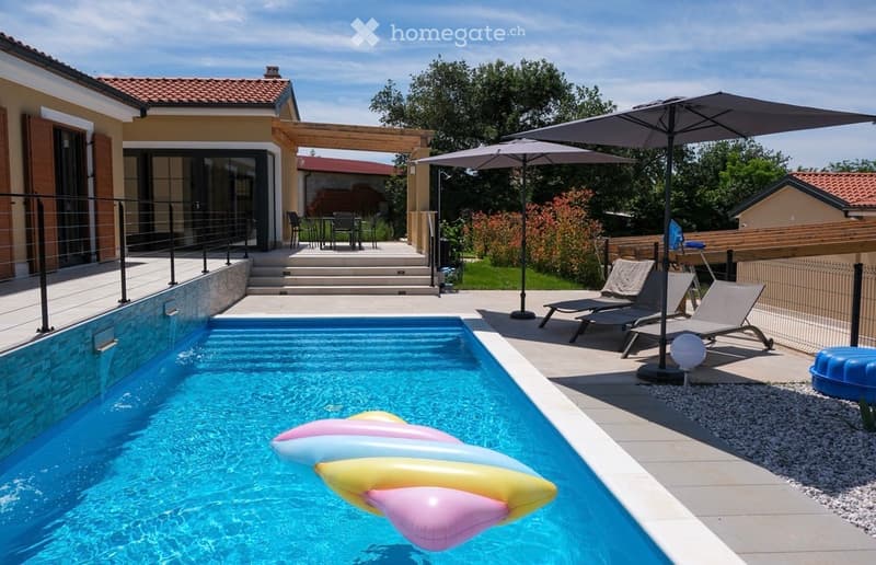 Outdoor area with a swimming pool, lounge chairs, umbrella, and a modern house in the background.