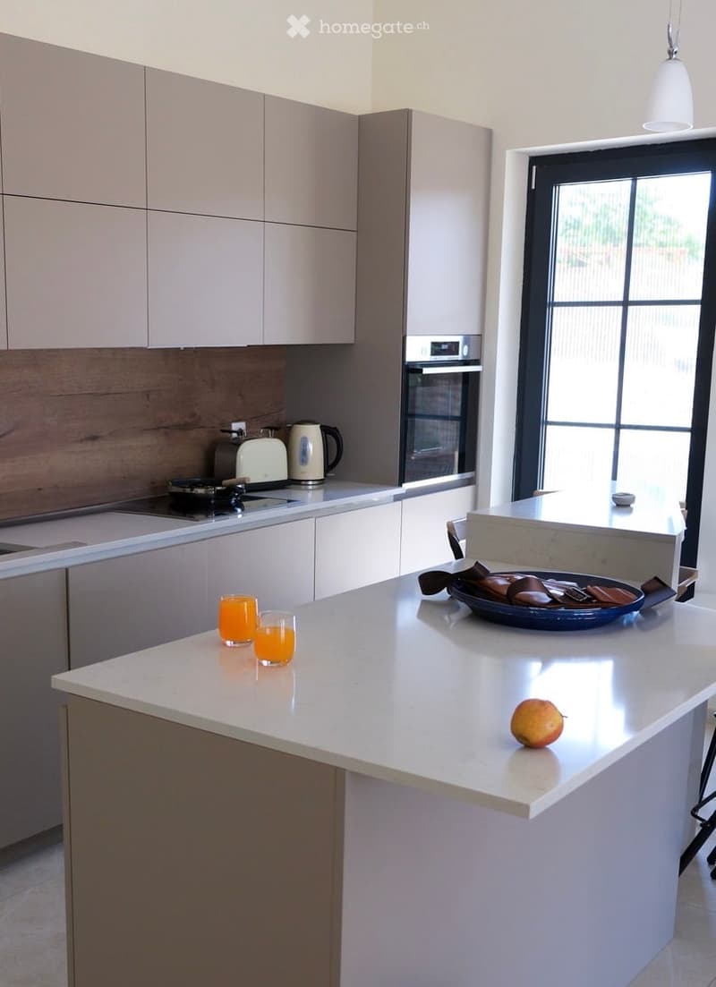 Modern kitchen with white cabinets, a central island, and various appliances including an oven, toaster, and coffee maker. The island features a bowl with snacks and a fruit, along with two glasses of orange juice.