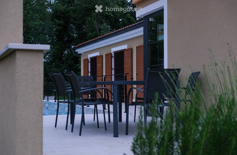 Outdoor patio area with a black metal table and four black chairs, adjacent to a swimming pool and a house with wooden shutters.