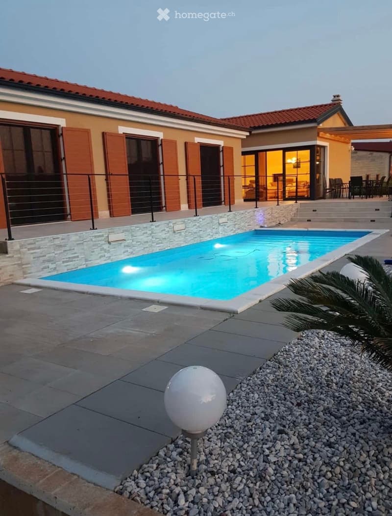 Modern outdoor area with a swimming pool, surrounded by a stone border, a palm tree, and a decorative spherical light fixture on a gravel bed.