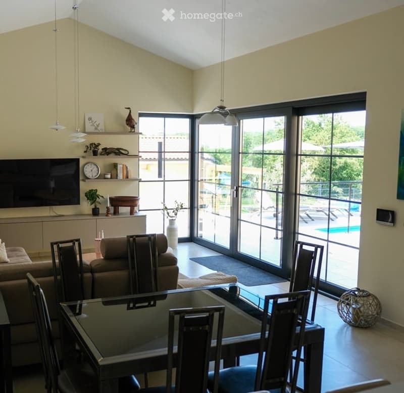 Bright living and dining area with large sliding glass doors opening to a pool area. Features a beige sofa, glass dining table with chairs, flat-screen TV, decorative shelf with plants and artwork, and modern lighting.