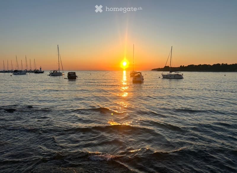 A serene sunset scene over a body of water with several sailboats and motorboats anchored, reflecting the warm hues of the setting sun.