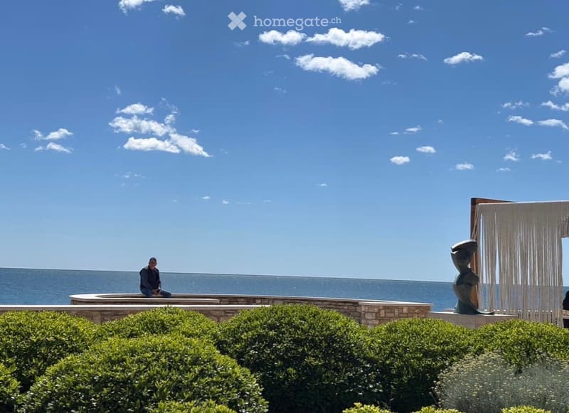 A serene outdoor area overlooking the ocean, featuring neatly trimmed hedges, a stone platform, and a modern sculpture with a draped fabric backdrop.