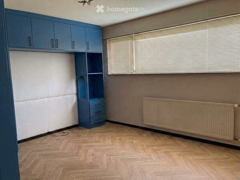 Empty room with wooden flooring, blue built-in storage unit, radiator, and window with blinds.