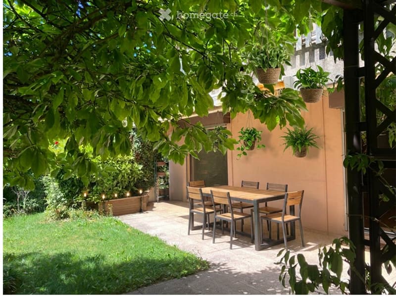 A shaded outdoor dining area with a wooden table and chairs, surrounded by greenery and hanging plants.