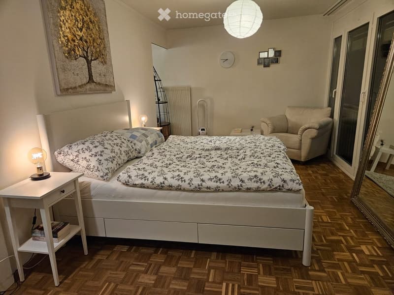 Cozy bedroom with a large bed, nightstand, armchair, and a large window with a view. The room features wooden flooring, a wall-mounted clock, and decorative artwork.