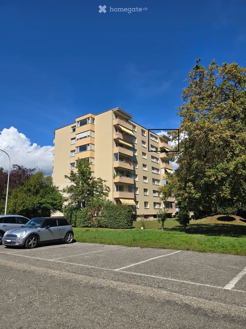 Multi-story residential building with balconies, surrounded by greenery and parked cars.