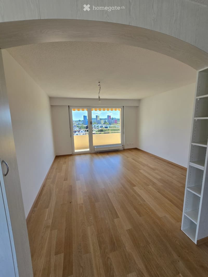 Empty room with wooden flooring, white walls, and a large window offering a city view. The room has a modern, minimalist design with a built-in shelf unit on one side.