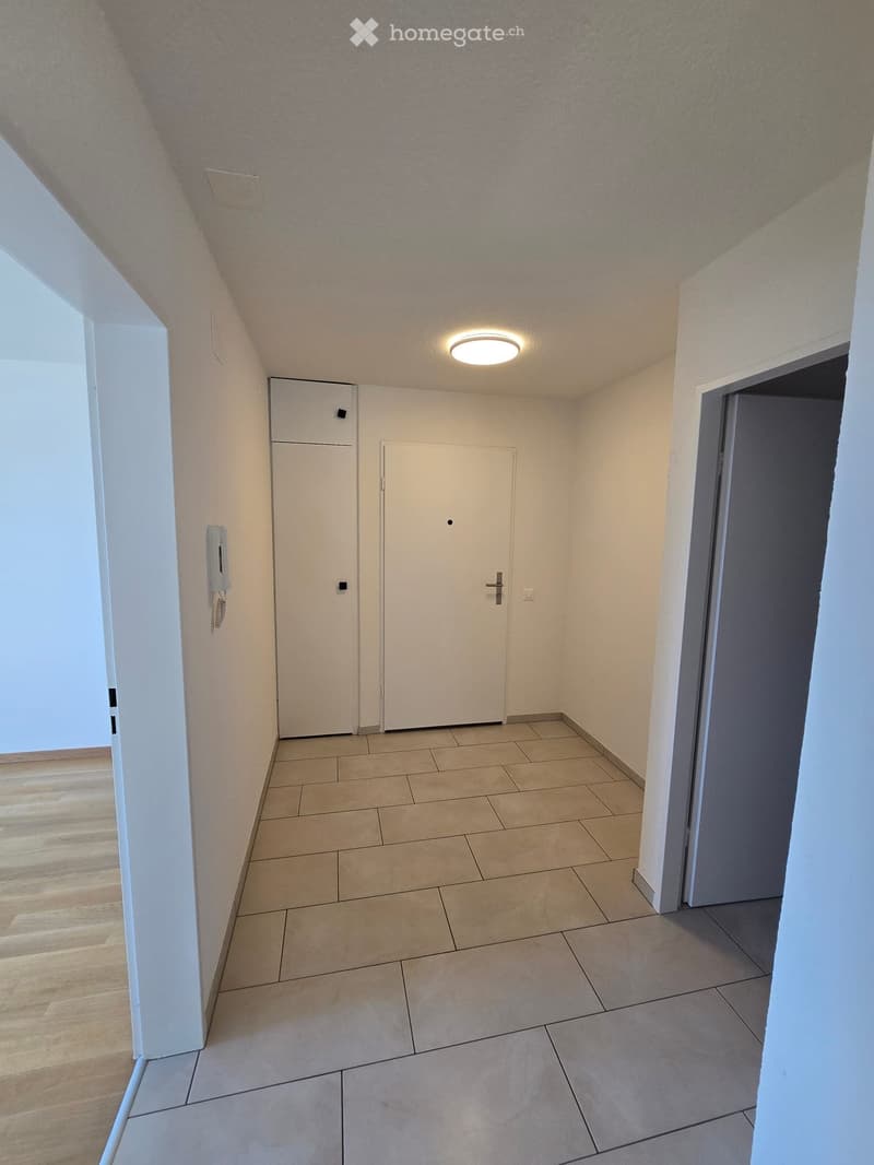 Empty hallway with tiled floor, wooden flooring on one side, white walls, and recessed lighting. Features two doors and a telephone on the wall.