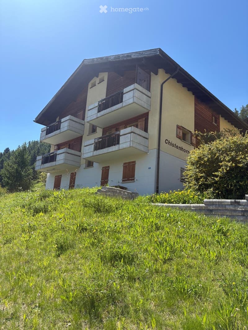Two-story house with balconies, wooden shutters, and a grassy slope in front.