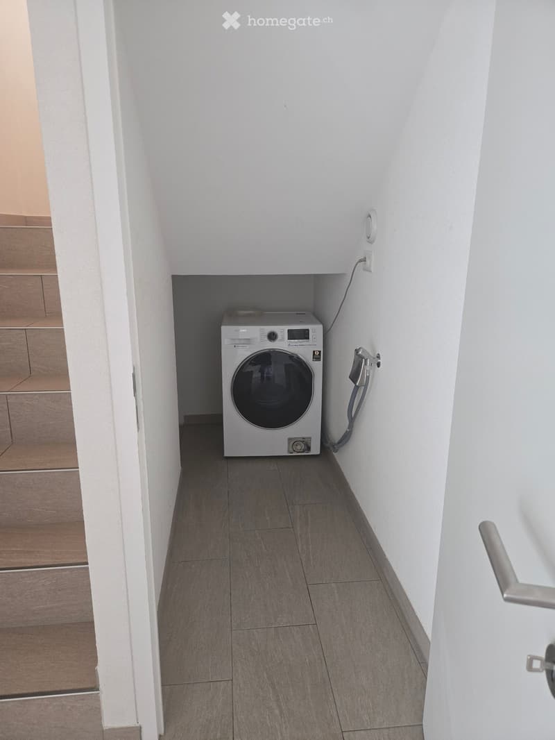 Narrow hallway with a private washing machine placed in a corner, white walls, grey tiled floor, and a visible electrical outlet.