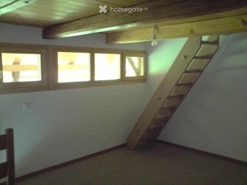 An attic space with exposed wooden beams, a wooden staircase leading up, and several windows letting in natural light. The room is empty with no furniture or furnishings.