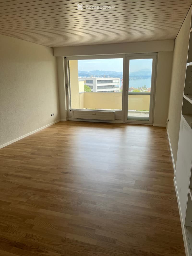Empty room with wooden flooring, large window with a view of the lake and mountains, radiator, and white walls.