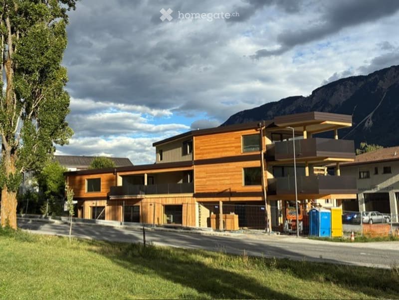 Modern multi-story building with wooden facade, balconies, and large windows, situated in a scenic area with mountains in the background.