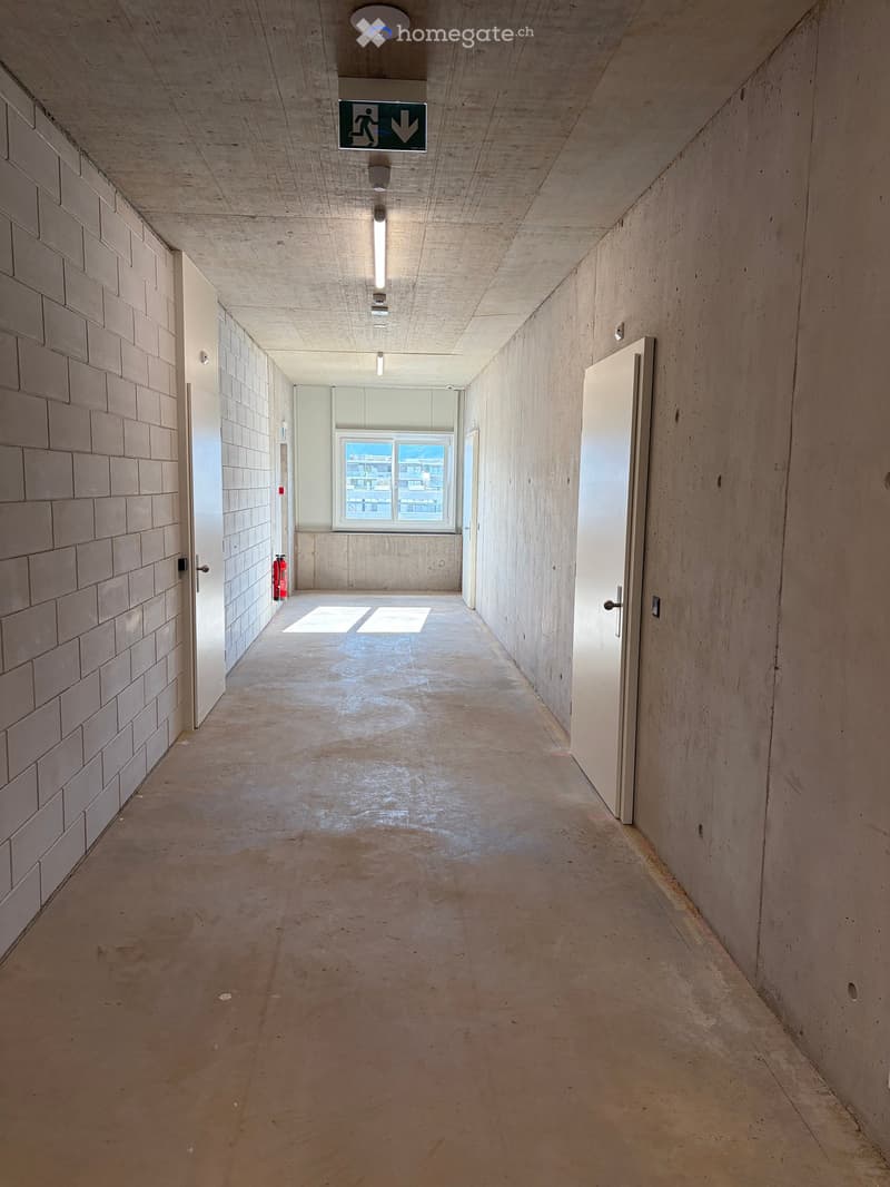 Empty hallway with concrete walls and floor, white doors, a window at the end, and a fire extinguisher on the left side.