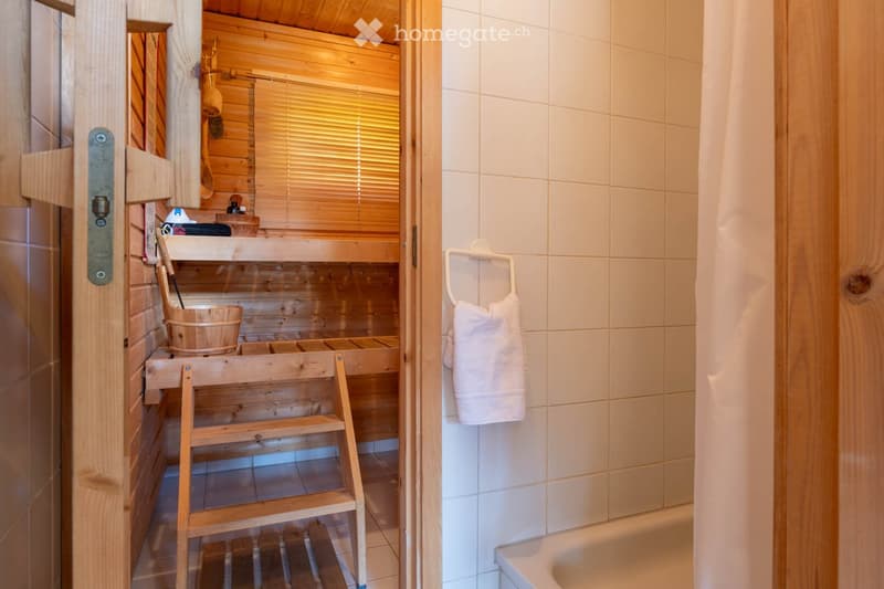 Bathroom. Bathroom with a wooden sauna area featuring a wooden bench, bucket, and ladder, adjacent to a tiled bathroom area with a bathtub and a towel hanging on a rack.