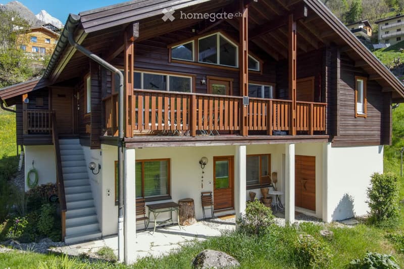 House. Two-story wooden house with a balcony, surrounded by greenery and mountains in the background.