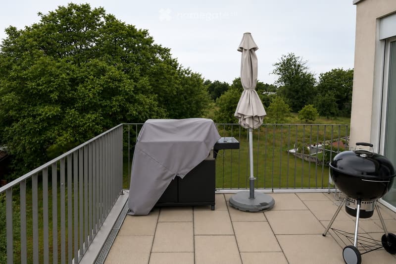 Balcony with a covered grill, a patio umbrella, and a railing overlooking a green landscape.