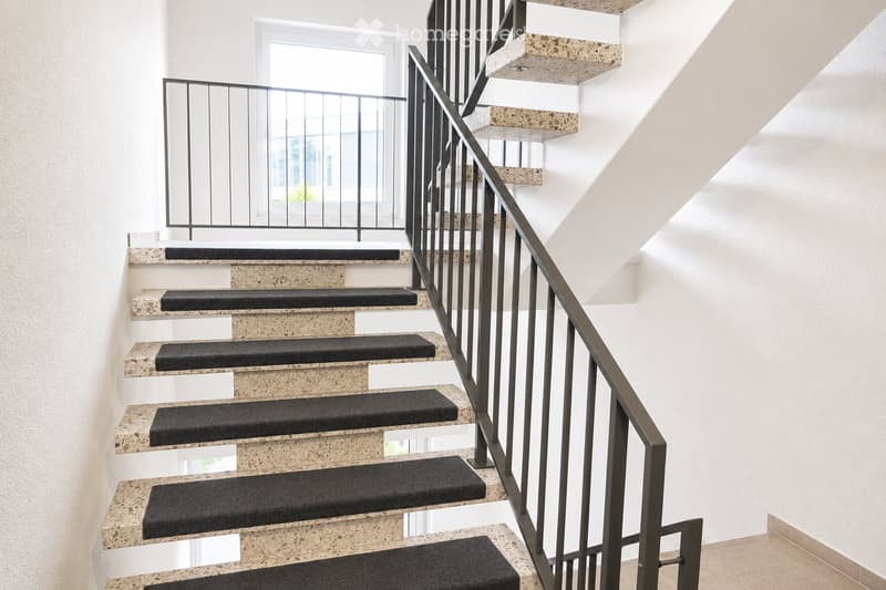 Modern staircase with beige stone steps and black carpet runners, featuring a sleek metal railing and a window at the top landing.