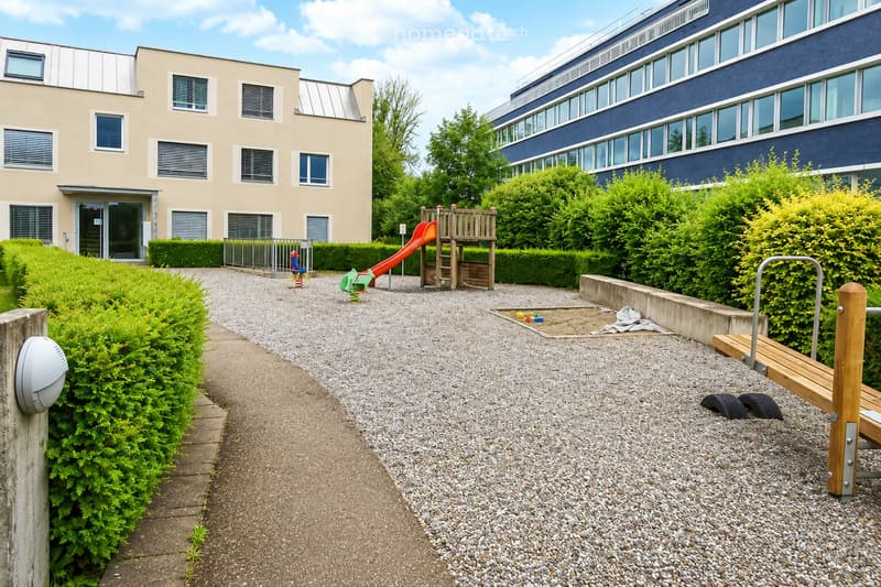 Outdoor playground area with a slide, climbing frame, and sandpit, surrounded by greenery and buildings.