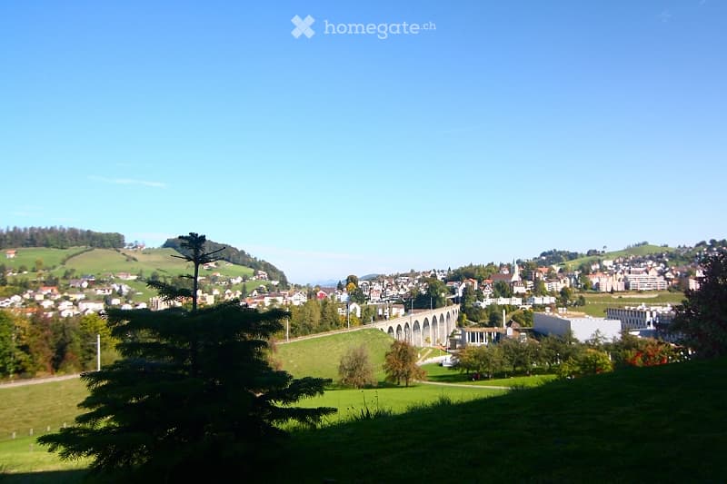 A panoramic view of a town with a mix of residential and commercial buildings, surrounded by green hills and a clear blue sky. A prominent aqueduct structure is visible in the distance.