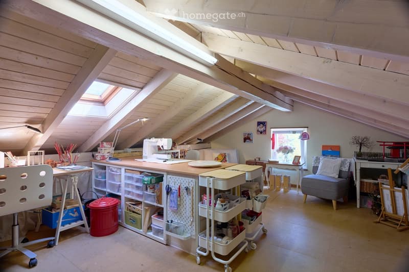 Brightly lit attic room with wooden beams and skylights, furnished with a large worktable, sewing machine, storage bins, a rolling cart, a sofa, and a chair.