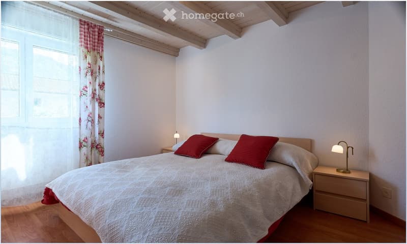 Cozy bedroom with wooden floor, white walls, and exposed wooden ceiling beams. Features a double bed with white bedding and red pillows, two nightstands with lamps, and floral curtains on the window.
