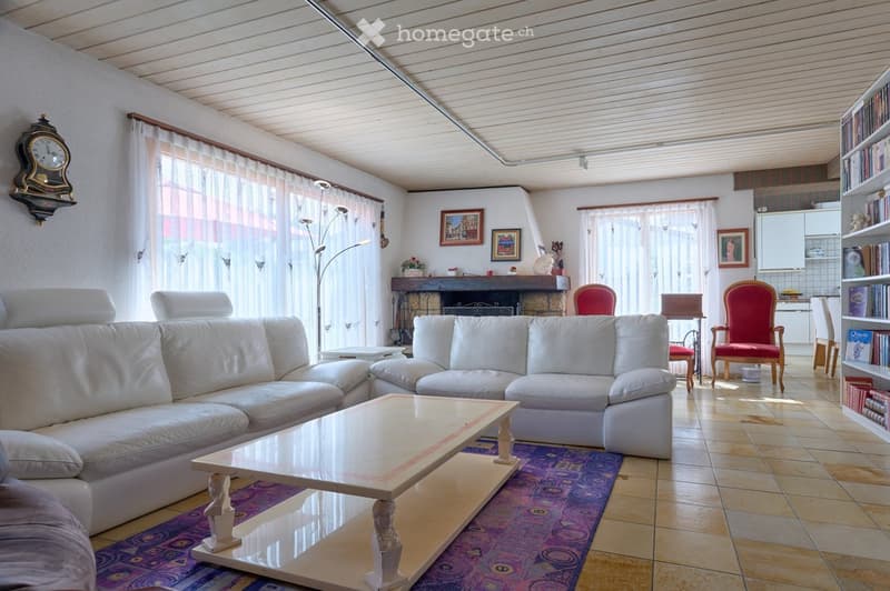 Bright living room with white leather sofas, a glass coffee table, a fireplace, and large windows with sheer curtains. The room features a tiled floor, a bookshelf, and a decorative clock on the wall.