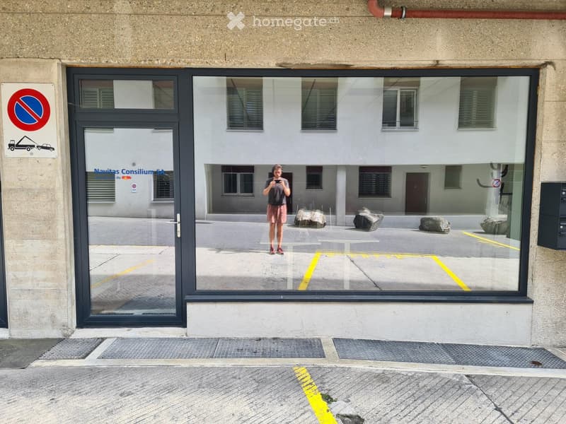 Building entrance with glass doors, a no-parking sign, and a reflection of a parking area with rocks and a person in the glass.