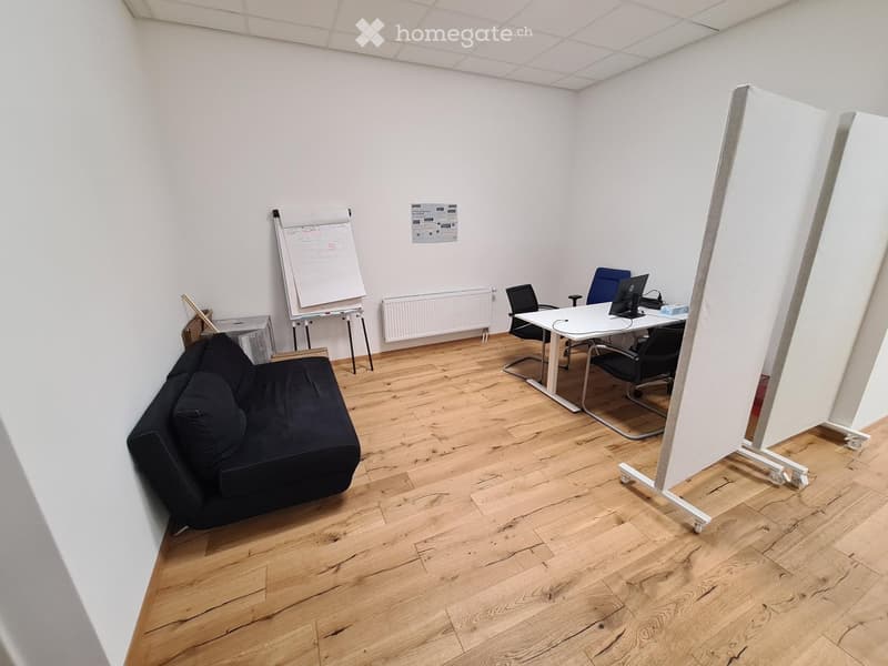 A modern, well-lit room with wooden flooring, a black sofa, a white table with chairs, a large screen, and a whiteboard on wheels.