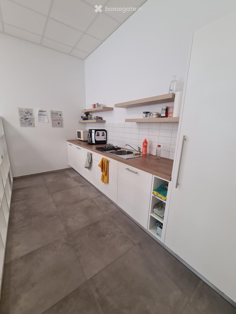 Modern kitchen with white cabinets, wooden countertop, and grey tiled floor. Includes microwave, coffee maker, sink, shelves, and various small items.