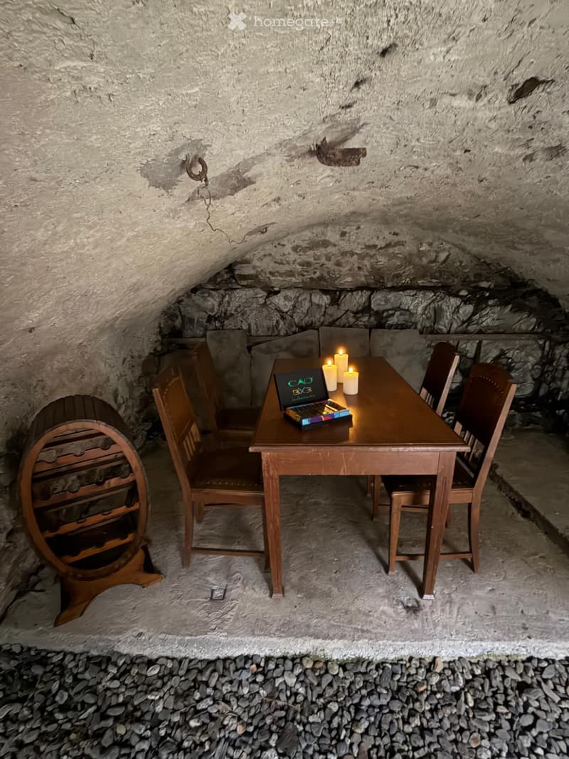 A rustic underground room with a wooden table, chairs, a laptop, candles, and a wine rack.