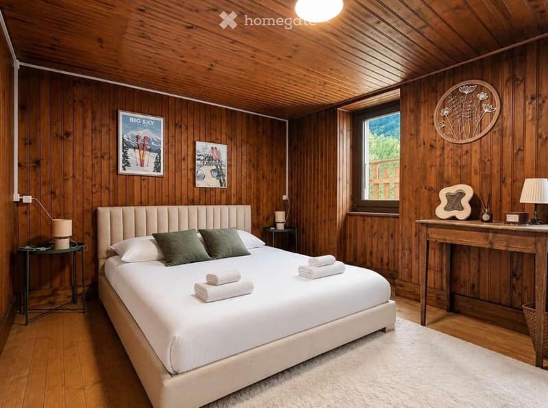 Cozy bedroom with wooden walls and ceiling, a large bed with white linens and green pillows, nightstands with lamps, a small table with a lamp, and decorative wall art.