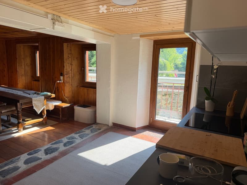 Bright, wooden-floored room with a dining table, chairs, a small stool, a cutting board, and a vase on a counter. Large windows offer a scenic view.